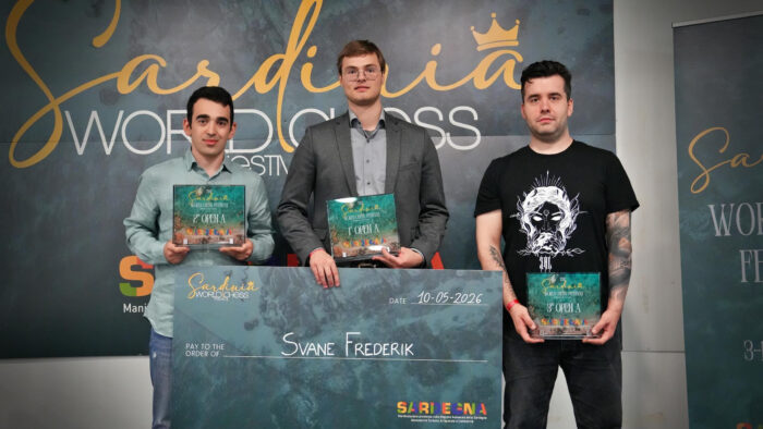 Церемония награждения шахматного турнира Sardinia World Chess Festival 2026. На фоне официального баннера стоят три призера группы Open A. В центре — победитель Фредерик Сване (Германия) в сером пиджаке, удерживающий большой призовой чек на свое имя и награду за 1-е место. Слева от него стоит Айк Мартиросян (Армения) в светло-зеленой рубашке с наградой за 2-е место. Справа — Ян Непомнящий (Россия) в черной футболке с графическим принтом и наградой за 3-е место. Все участники держат в руках фирменные стеклянные призы фестиваля (Сардиния, 10 мая 2026)